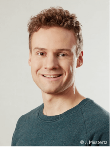 Jakob Lindenthal lächelnd, mit grünem Pullover vor hellem Hintergrund.
